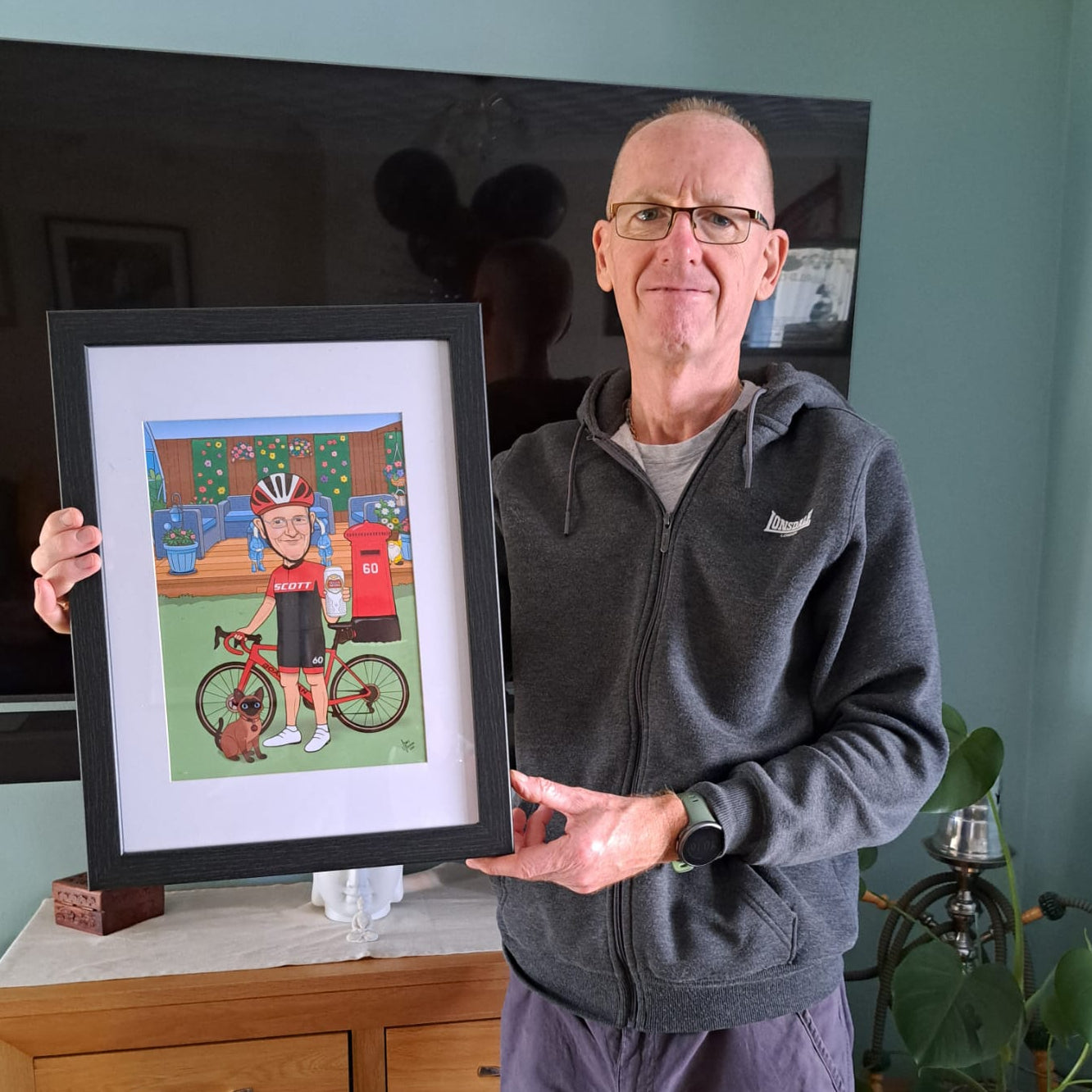 Man holding a framed picture of a cyclist caricature in a living room.