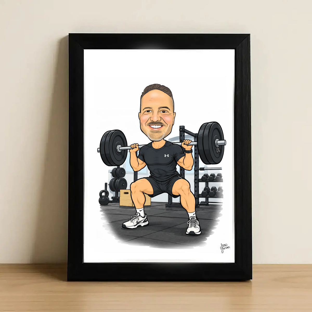 A framed caricature art print of a man lifting a barbell in a gym, shown leaning against a wall on a wooden surface. The print features a man in a black Under Armour t-shirt and shorts with white sneakers, set against a white background inside a black frame.